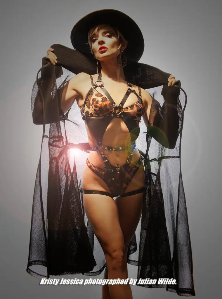 fashion model posing in strappy leopard print lingerie with long see through trench coat, sassy glasses and a big black hat shot in a studio with a white background and a starbust light coming from behind her