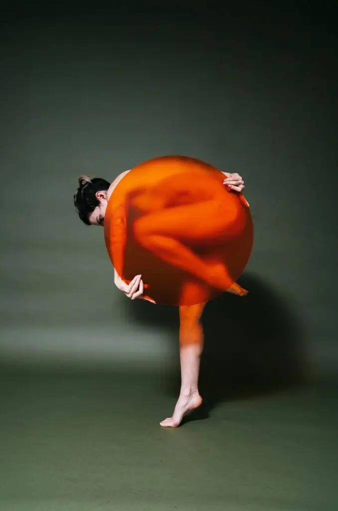 Unique creative studio photo of nude model in angular standing pose with one leg bent and posing arching downward with orange semi-transparent circle obscuring her body on a green paper backdrop