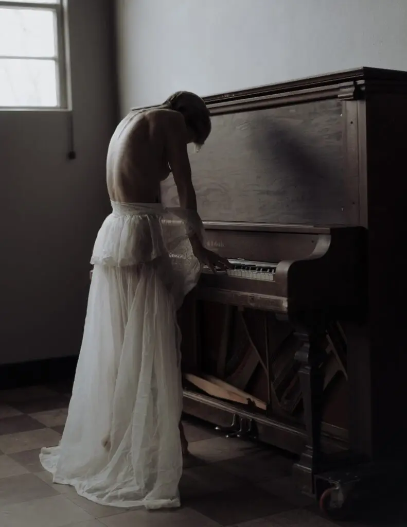 creepy dark photo of partially nude model in sheer vintage gown playing an abandoned piano with back arched upwards
