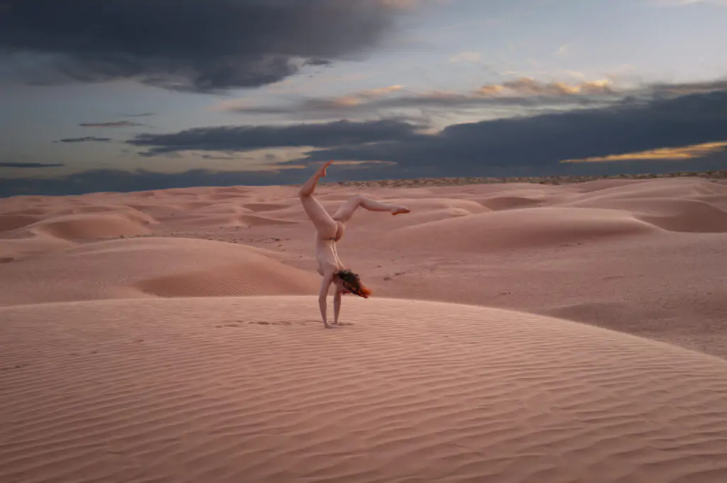 Model in sand dunes with dramatic sky doing a handstand nude
