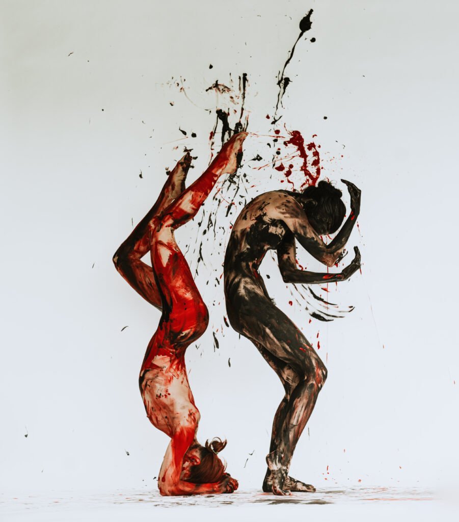 Two slender models posing vertically in odd angular positions with red and black paint splattered all over their bodies, against a white background with some paint splashes on it