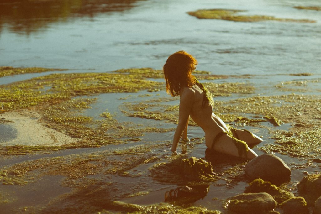 Pale woman crouched in a river with algae and seaweed all around and partially covering her body in the sunset