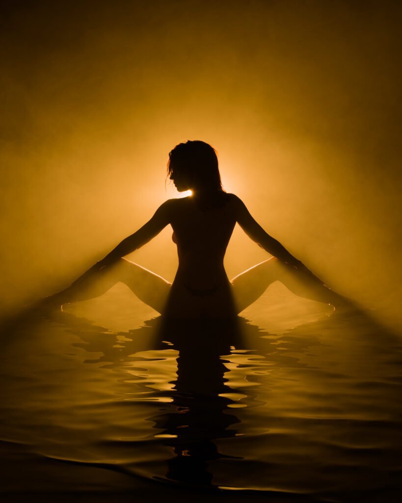 Model making shapes with arms and legs sitting in shallow water with golden amber foggy light silhouetting her