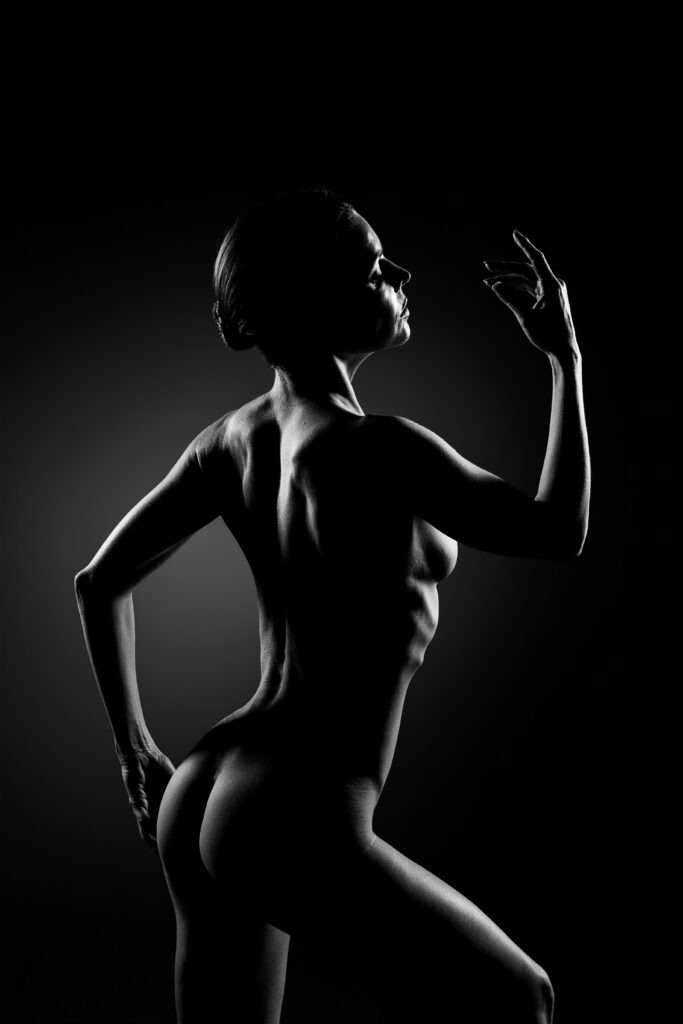 Dark studio photography of nude model posing from the back showing definition in arms, glutes, ribs and back with side lit aesthetic features, artistic