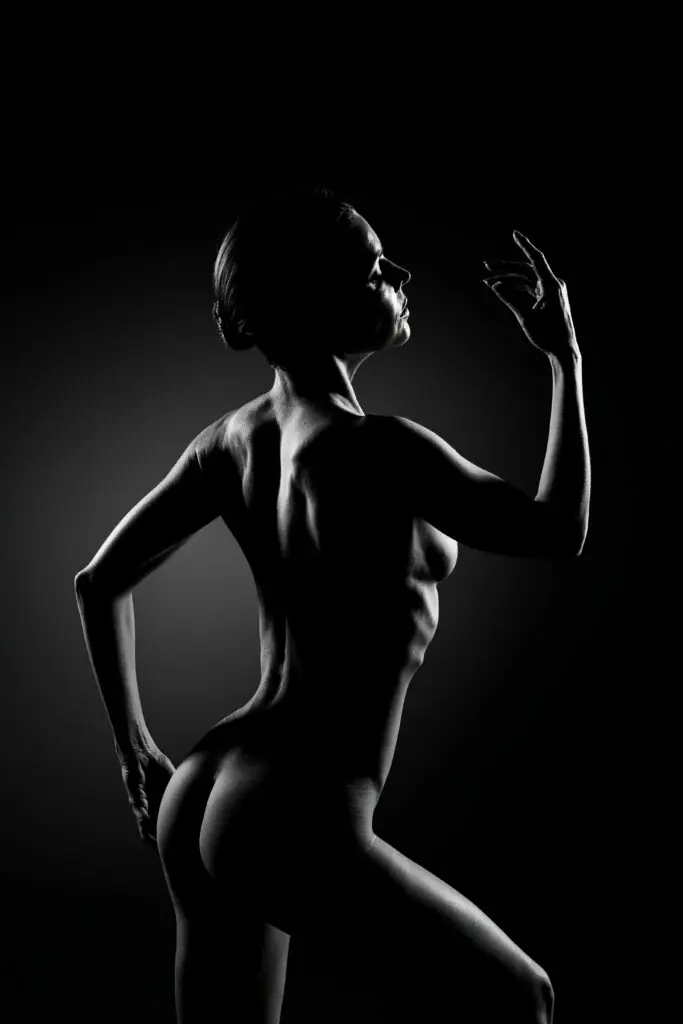 Dark studio photography of nude model posing from the back showing definition in arms, glutes, ribs and back with side lit aesthetic features, artistic