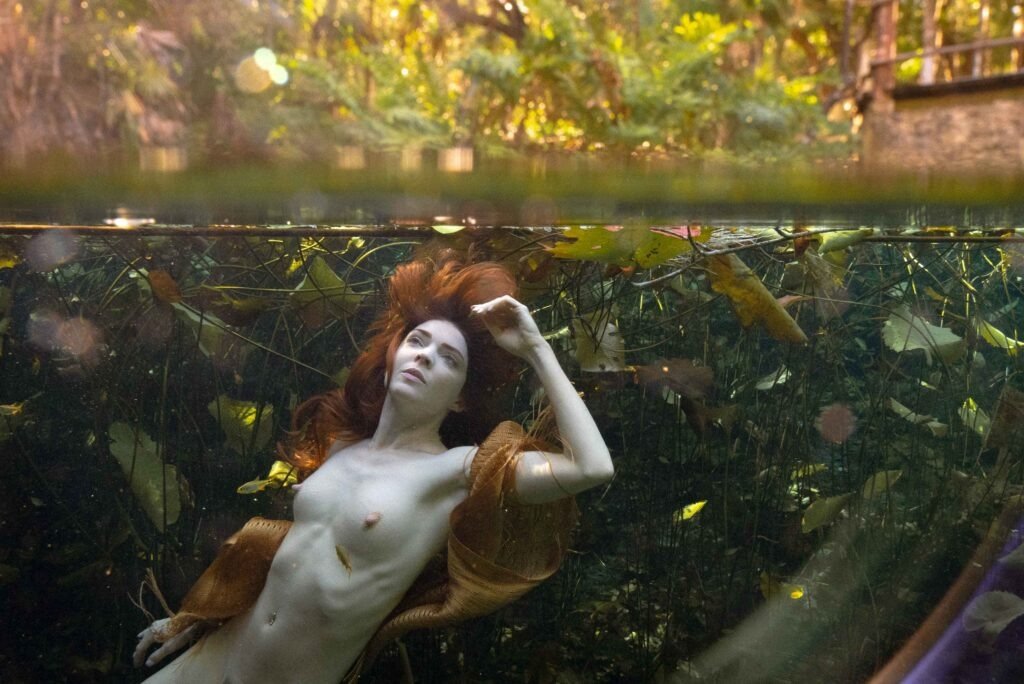 fine art underwater nude image of pale slender model Kristy Jessica with long red hair posing in a spring with small fish and lily pads all around