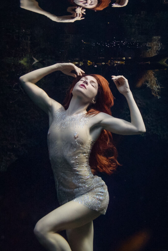 Elegant slender pale model posing underwater in crystal bodycon dress, her face just below the surface