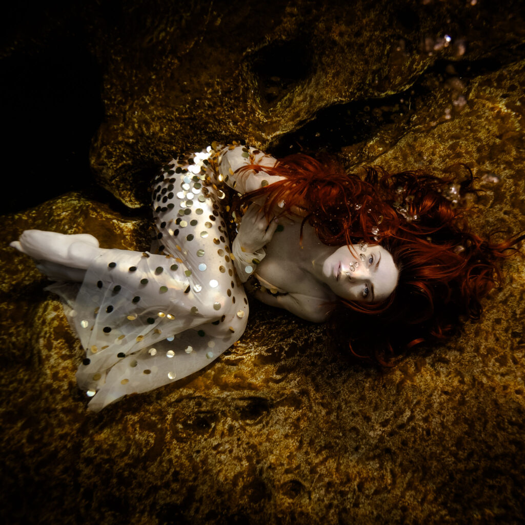Elegant model in gold shiny dress and red hair curled up underwater on yellow textured rocks