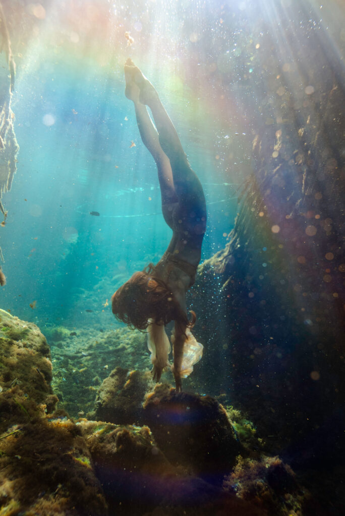 Model diving down into a crystal blue spring with rainbow light leak and green mossy rocks