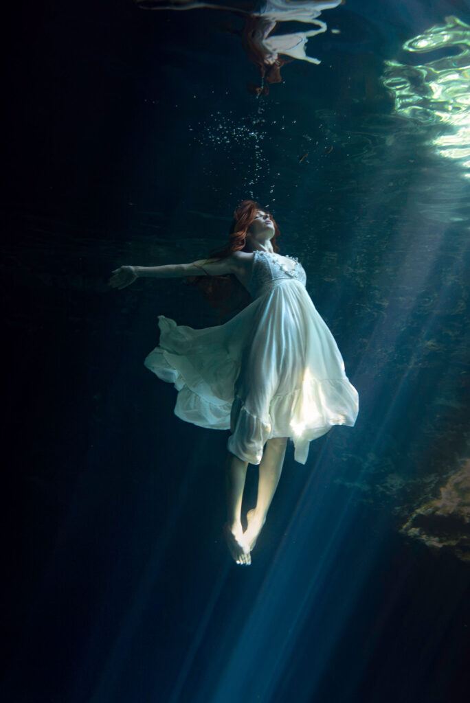 beautiful underwater photo of a model in a pale dress dancing in beams of light against a dark water spring background